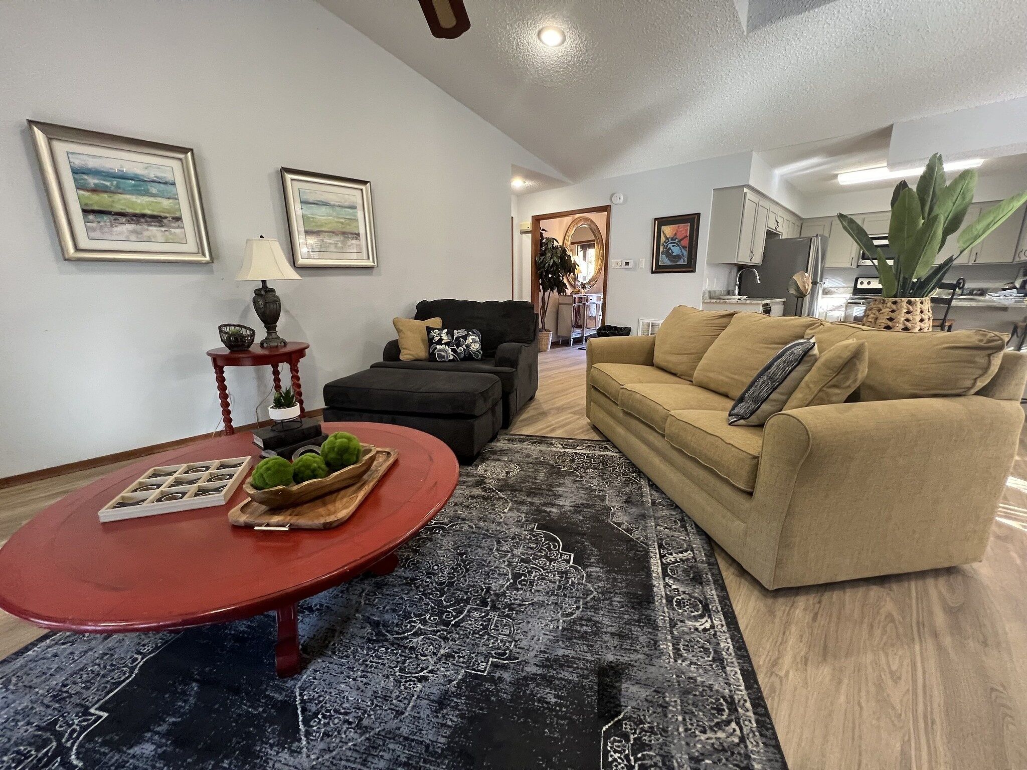 Una sala de estar con una mesa de centro roja, un sofá beige y un sillón negro. Hay dos cuadros enmarcados en la pared, una lámpara sobre una mesita auxiliar y una zona de cocina al fondo.