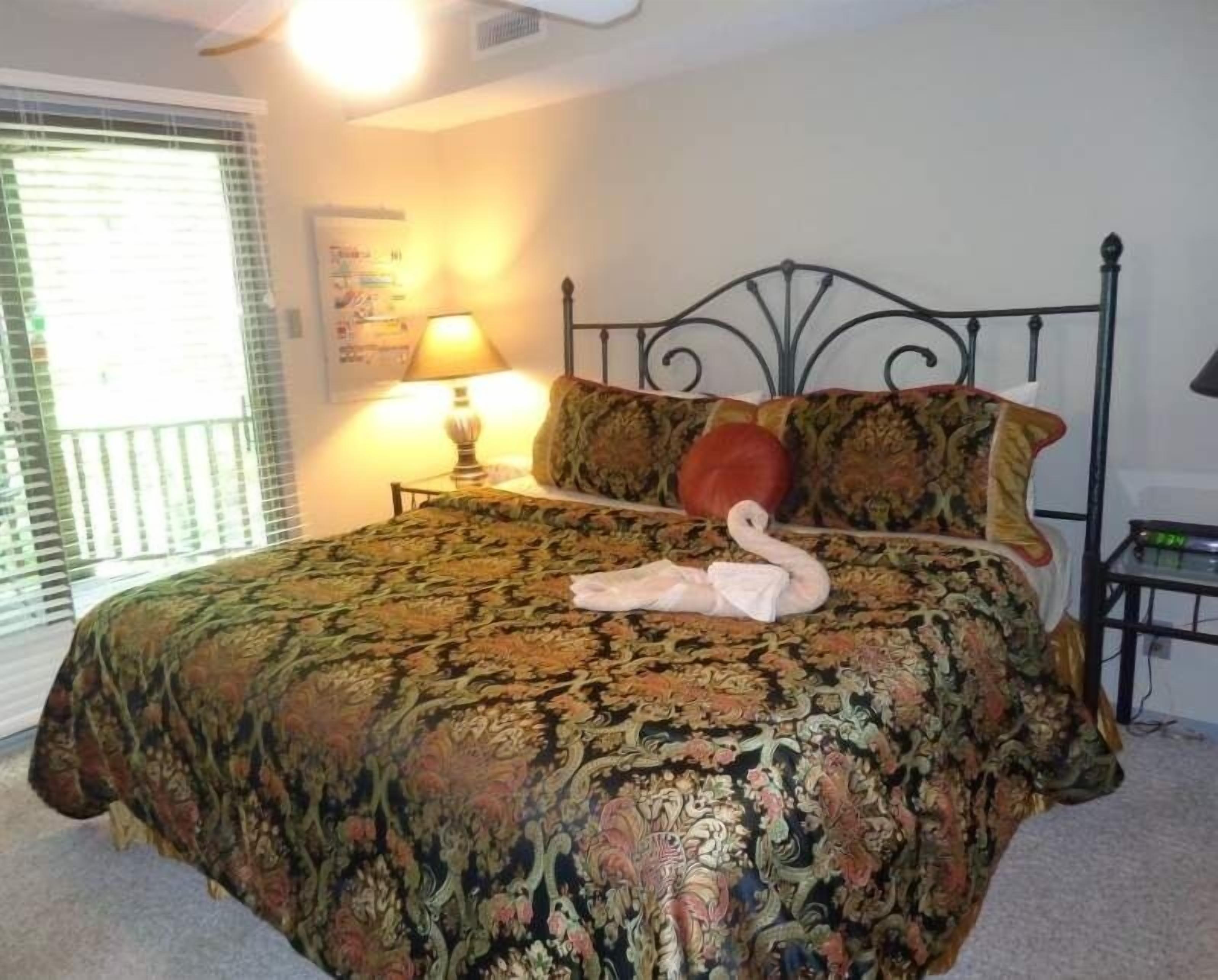 A bedroom with a patterned bedspread, a lamp on the nightstand, and a window with blinds.