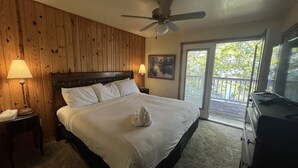 A bedroom with a wooden headboard, a bed with white linens, a ceiling fan, a nightstand with a lamp, a dresser, and a view of the outdoors through a glass door. - Lake view two bedroom, two bath log-sided, luxury Harbor North loft cottage with hot tub overlooking Lake Ouachita. by RedAwning (Mount Ida)