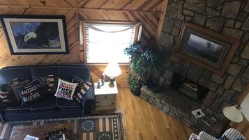 A cozy living room with a wooden ceiling, a stone fireplace, a couch with patriotic pillows, and a view of the outside.