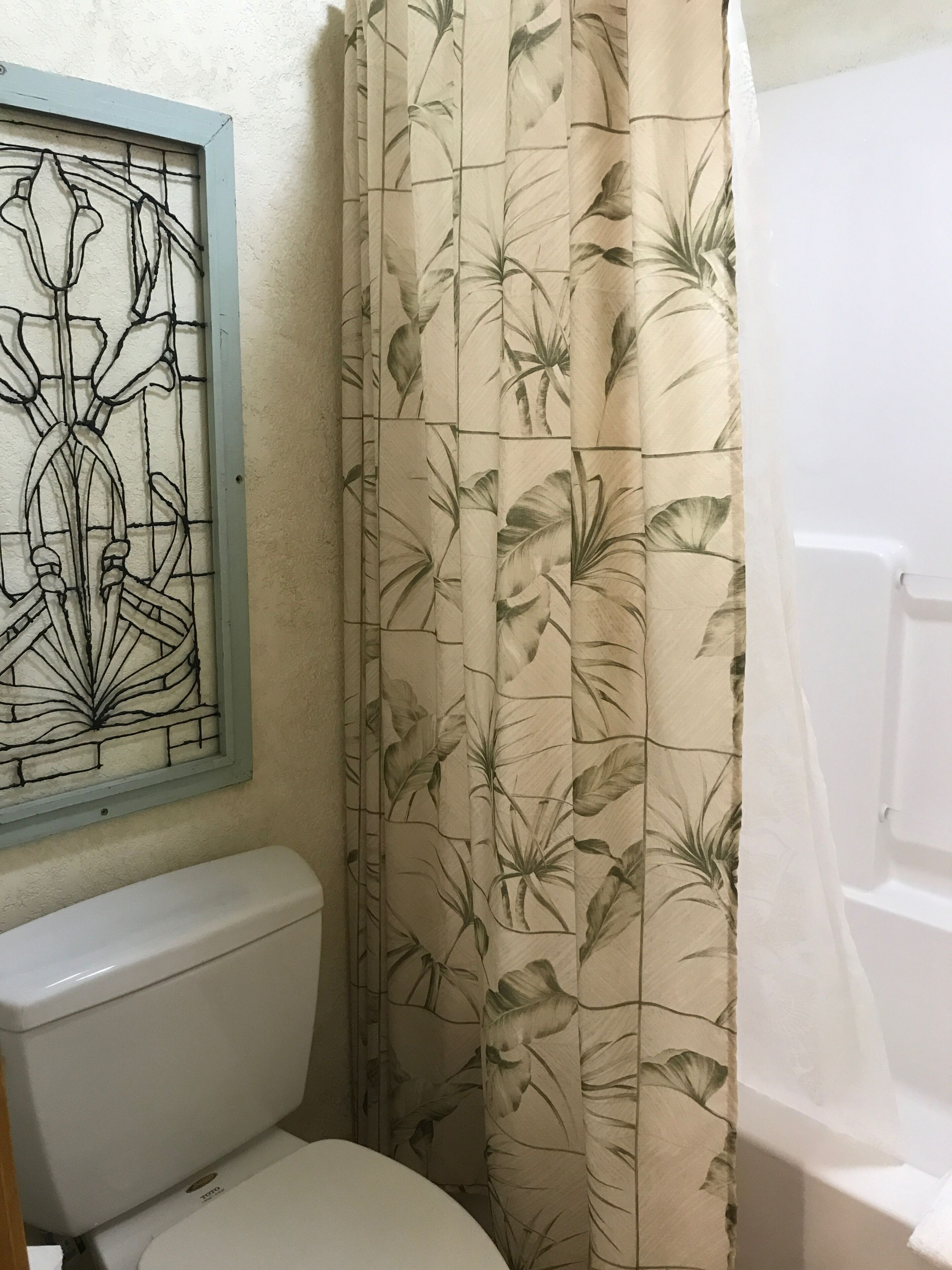 A bathroom with a toilet, a shower curtain with a floral pattern, and a stained glass window above the toilet.