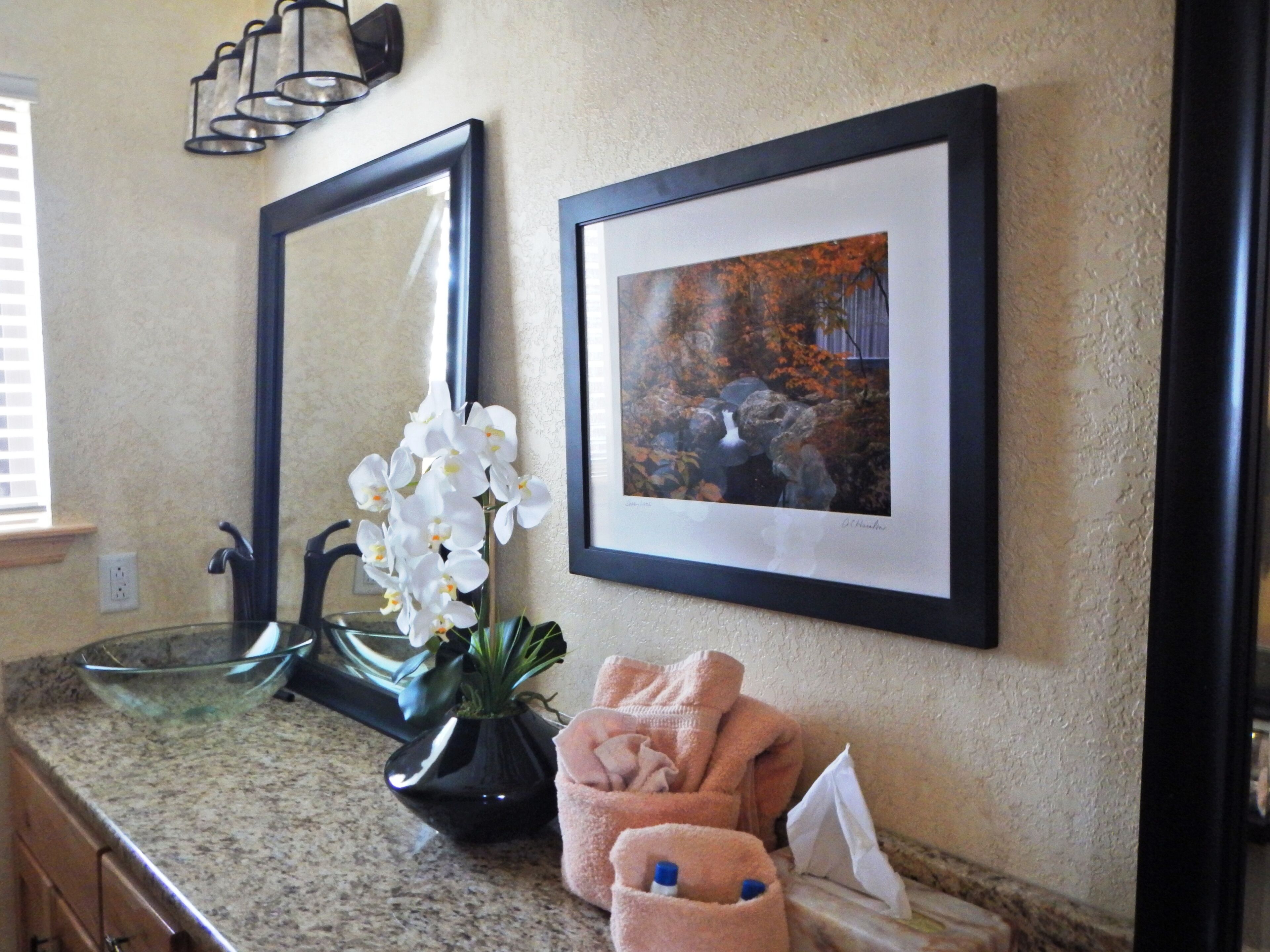A bathroom with a granite countertop, a black vase with white orchids, a framed picture of a waterfall, and a towel rack with a pink towel.