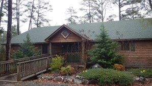 A log cabin with a porch, surrounded by trees and a gravel driveway. - Lake view three bedroom, three bath cottage with private hot tub overlooking Lake Ouachita. by RedAwning (Mount Ida)
