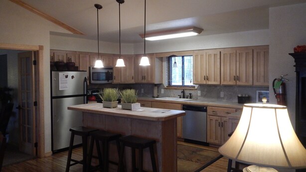 A kitchen with wooden cabinets, a stainless steel refrigerator, a microwave, a sink, and a small island with stools. There are pendant lights above the island and a table lamp on the floor. - Lake view three bedroom, three bath cottage with private hot tub overlooking Lake Ouachita. by RedAwning (Mount Ida)