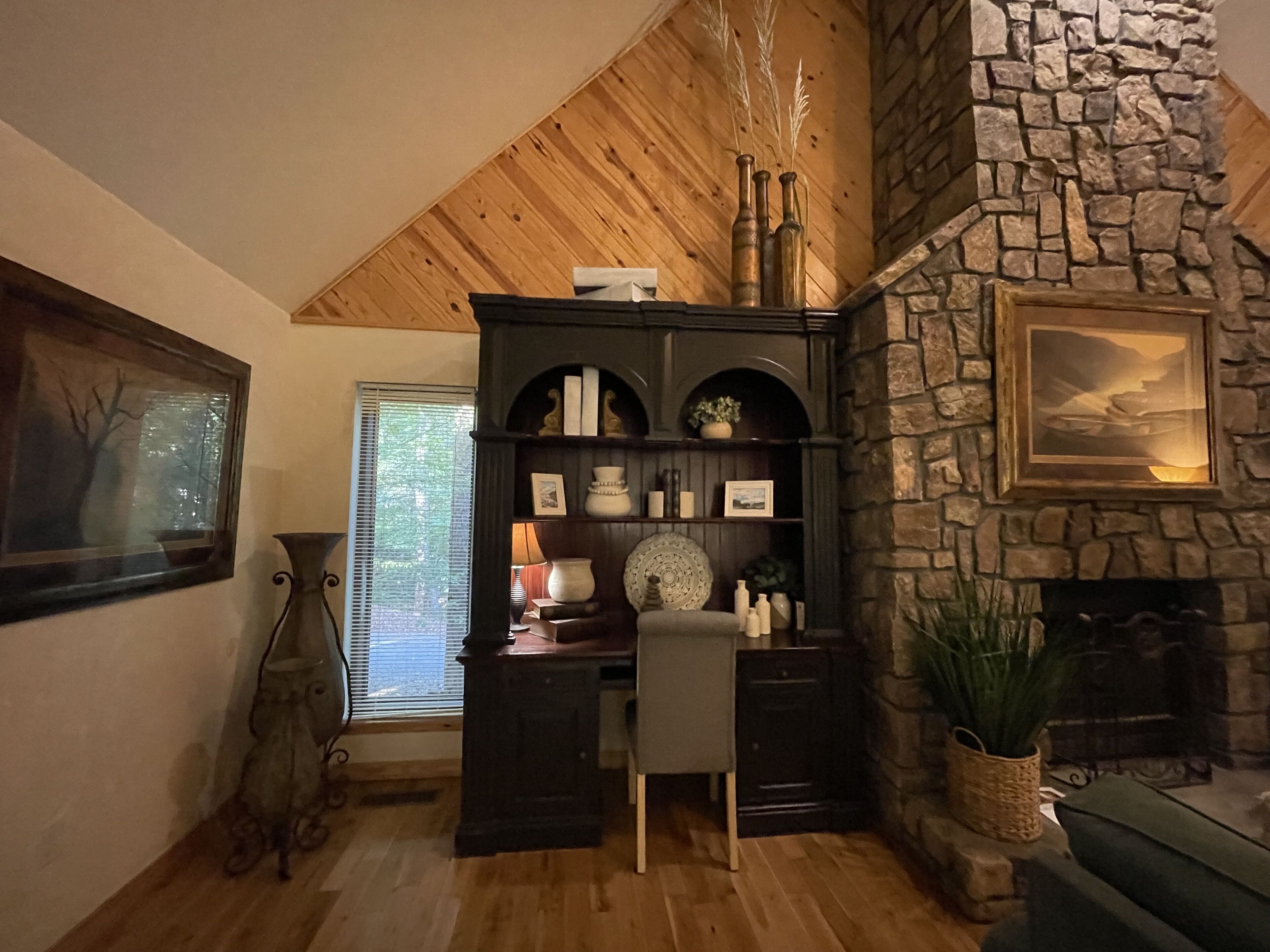 Uma sala de estar aconchegante com lareira de pedra, teto de madeira e uma estante escura de madeira repleta de livros e objetos decorativos.