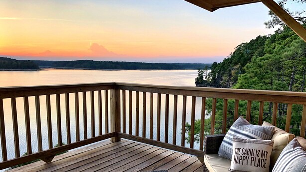 A wooden deck with a view of a lake at sunset, a table, and wicker furniture with pillows. - Magnificent Four Bedroom, Two 1/2 Bath luxury, log-sided Harbor North cottage with incredible views of Lake Ouachita. by RedAwning (Mount Ida)