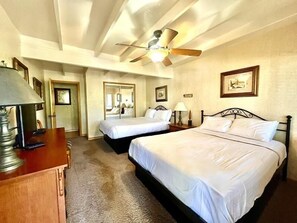 Quarto de hotel com duas camas, um ventilador de teto, uma escrivaninha com luminária e quadros nas paredes.