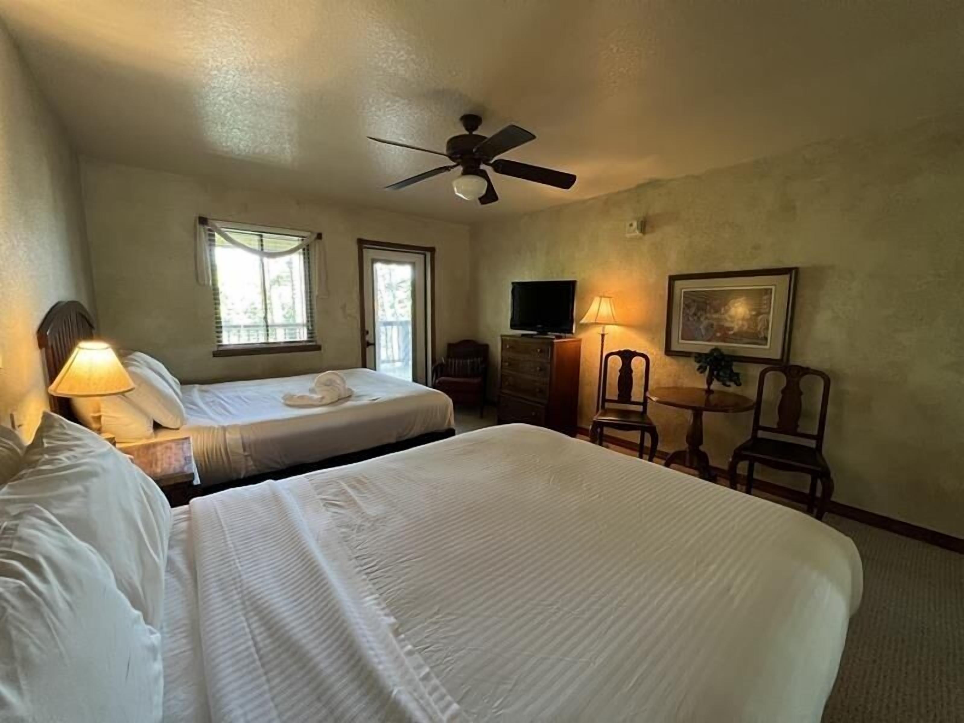 Une chambre d’hôtel avec deux lits, un ventilateur de plafond, une télévision, un bureau avec une chaise et un tableau au mur.