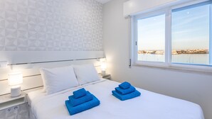A modern hotel room with a large bed, white bedding, and a view of the sea through a large window.