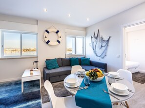 A modern living room with a sofa, dining table set for four, and a view of the city through large windows. - Torri riverview apartment (Portimão)