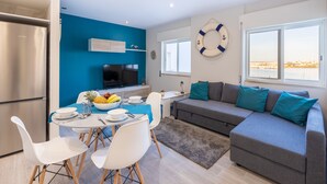 A modern living room with a grey sofa, teal cushions, a dining area with white chairs and a table, a flat-screen TV, and a large window with a view.