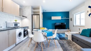A modern kitchen with a washing machine, a dining area with a blue tablecloth, and a living area with a TV and sofa.