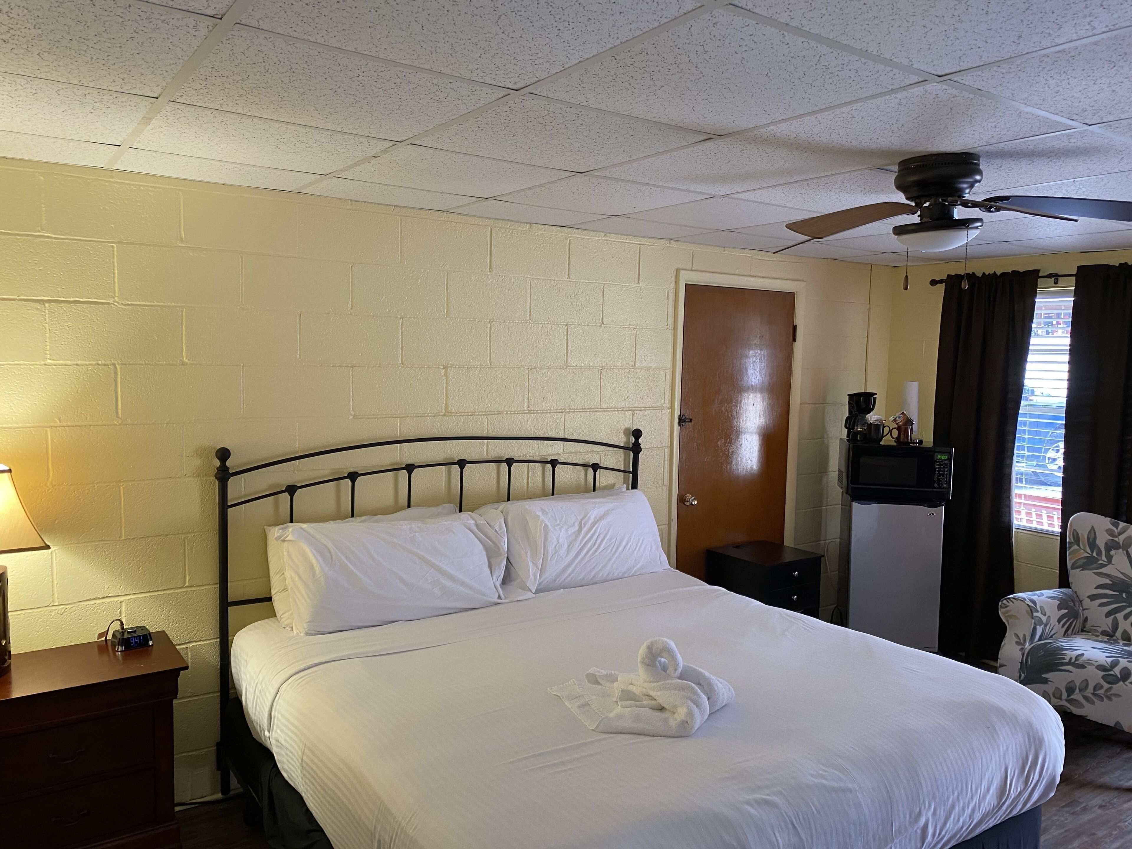 Hotelkamer met een bed, nachtkastje, bureau met lamp, stoel, plafondventilator, deur en raam met gordijnen.