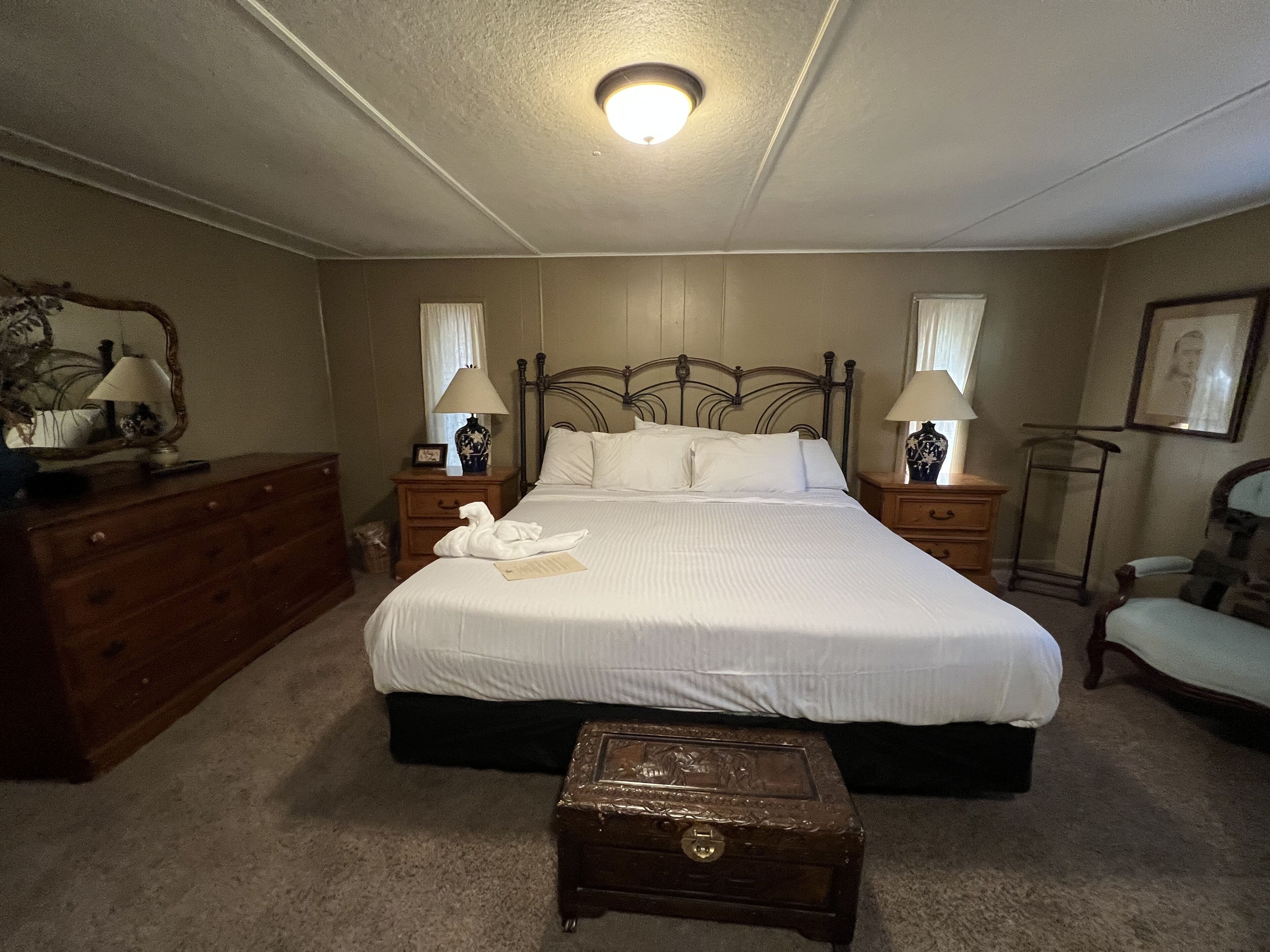 A neatly arranged bedroom with a large bed, two bedside tables with lamps, a dresser, a small wooden chest, and a chair.