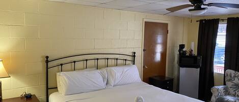 A hotel room with a bed, a nightstand, a desk with a lamp, a chair, a ceiling fan, a door, and a window with curtains.