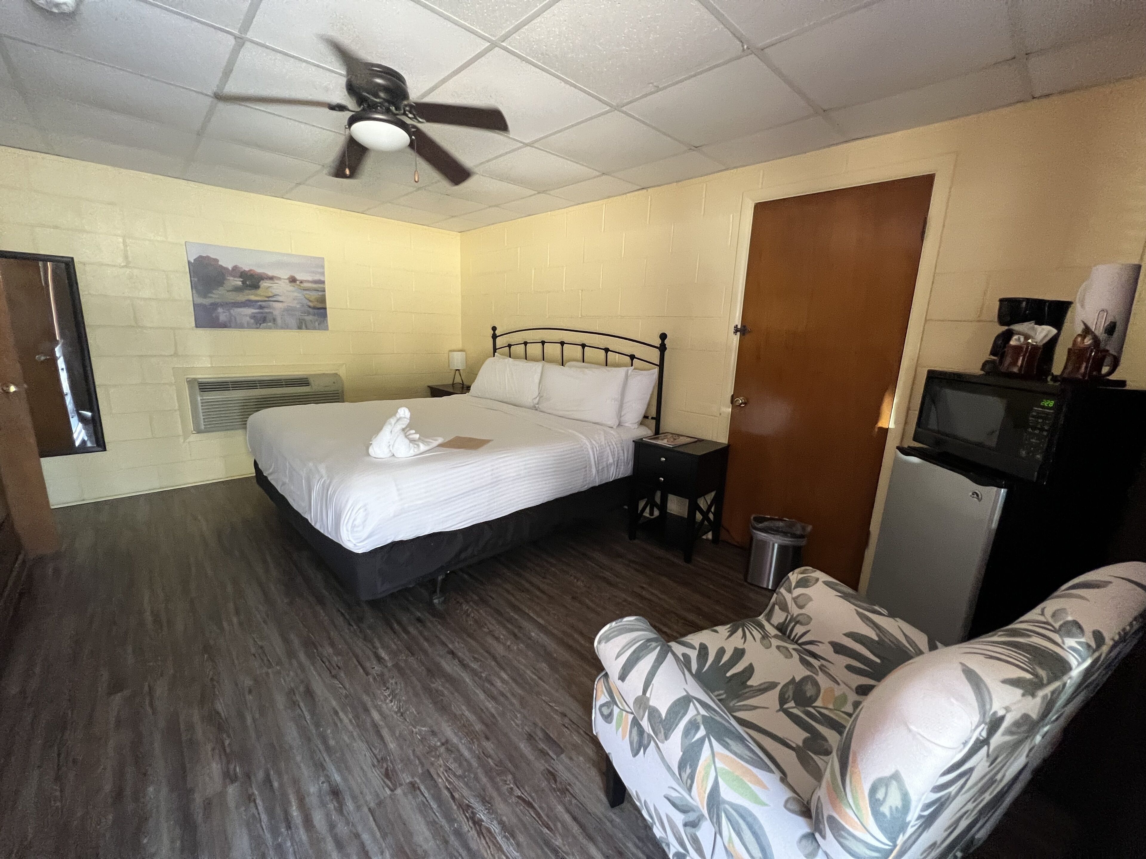 A hotel room with a bed, a chair, a ceiling fan, a microwave, and a trash can.
