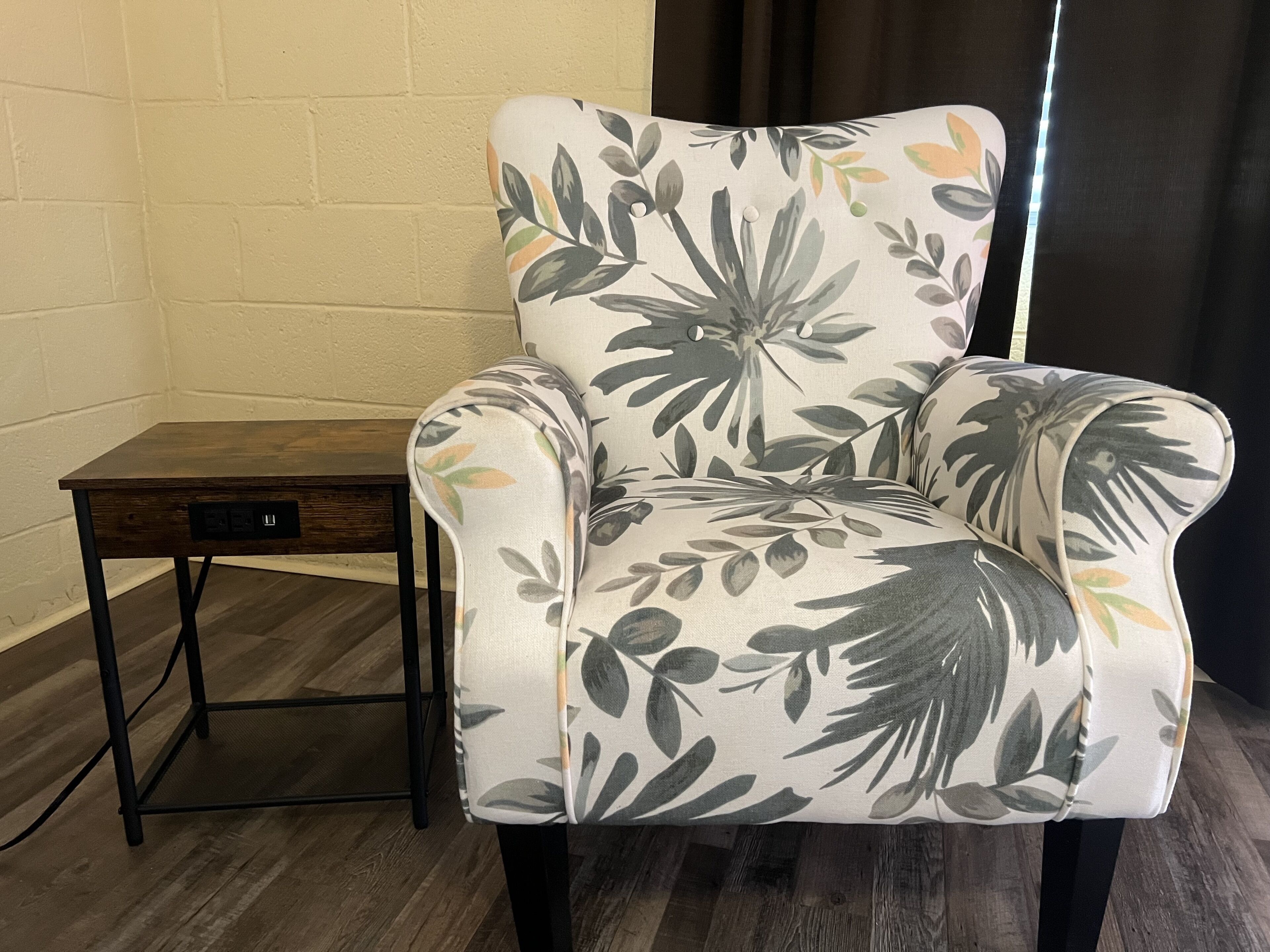 A chair with a floral pattern, a small table with a drawer, and a wooden floor.