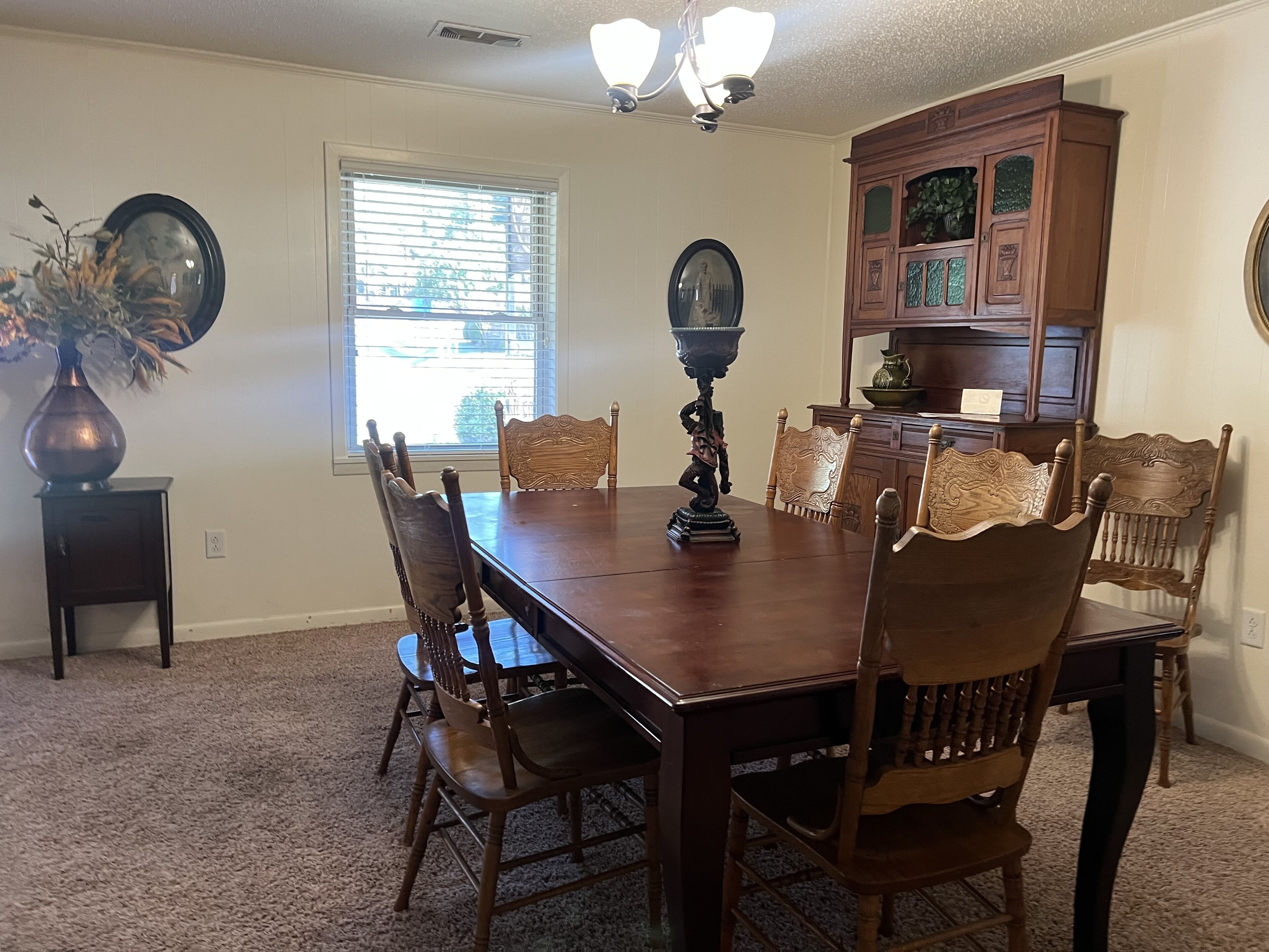 A dining room with a wooden table, six chairs, a vase with dried flowers, a mirror, and a cabinet with glass doors.