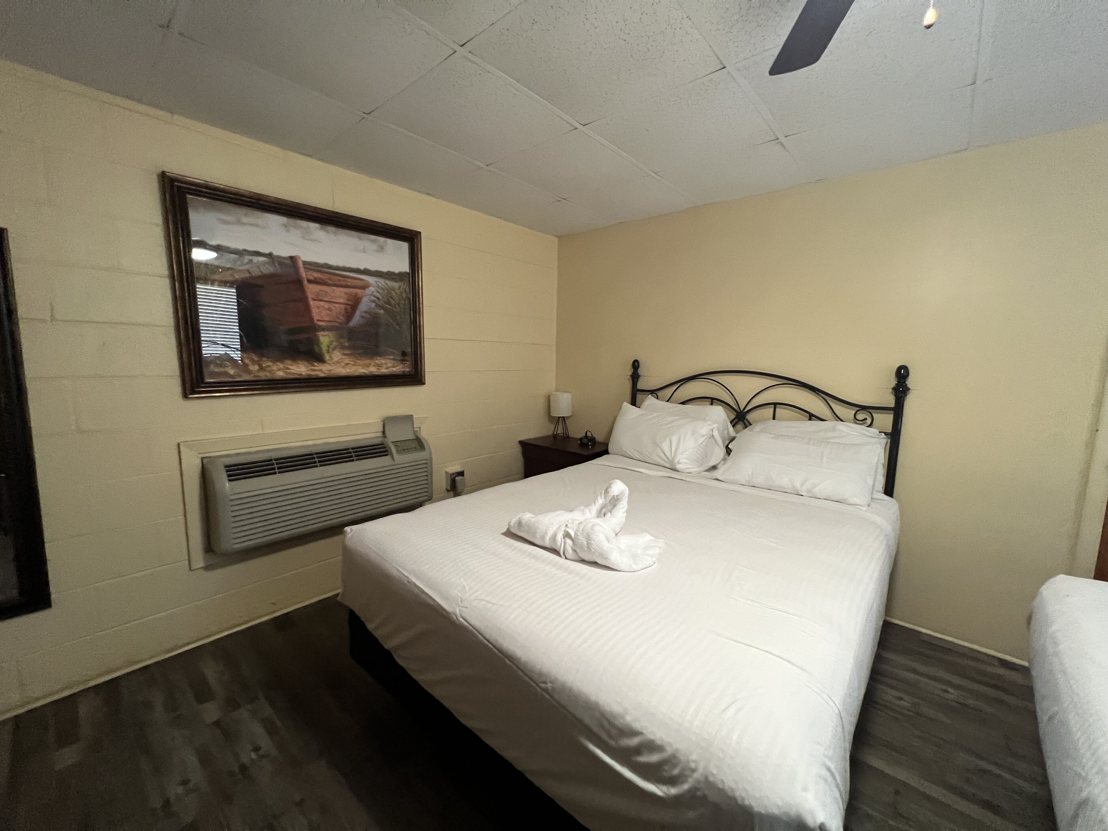 A hotel room with a bed, a framed picture on the wall, a heater, and a nightstand with a lamp.