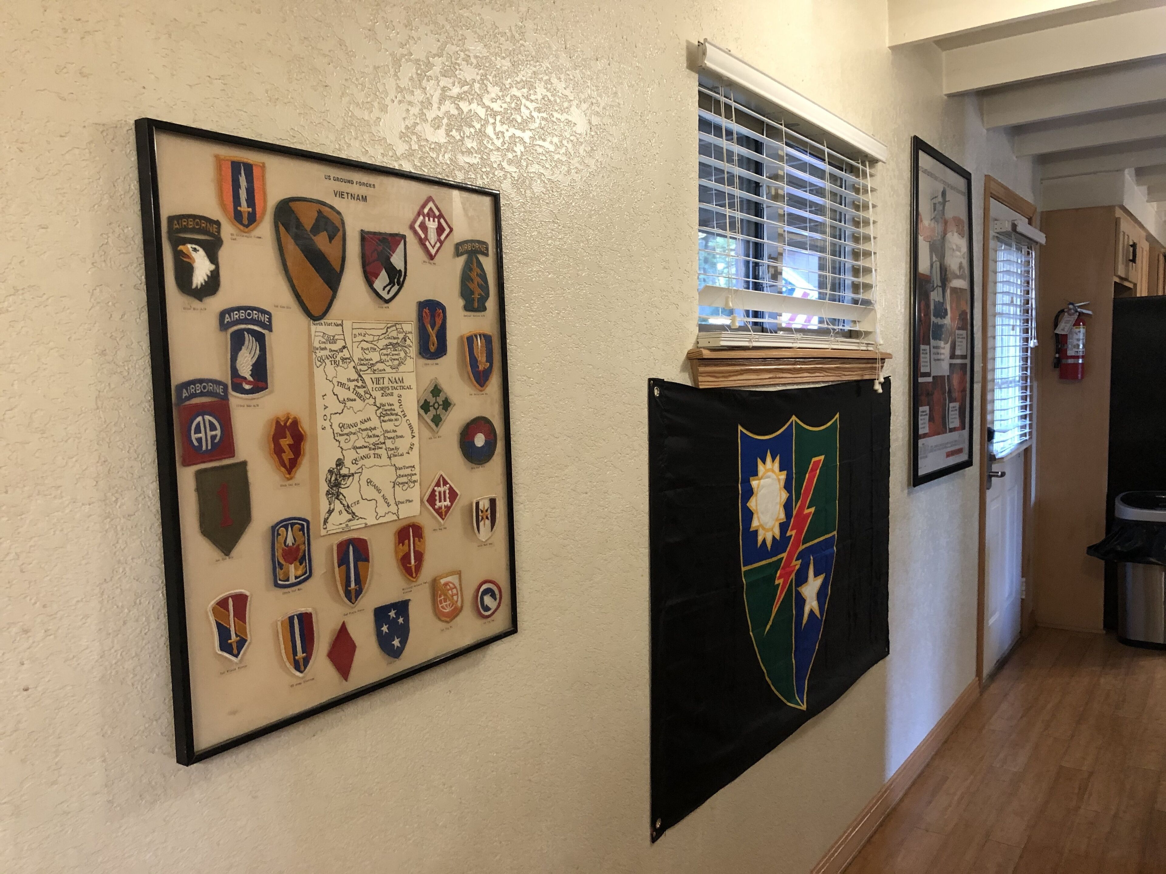A hallway with a framed collection of military unit patches on the left and a banner with a crest on the right.