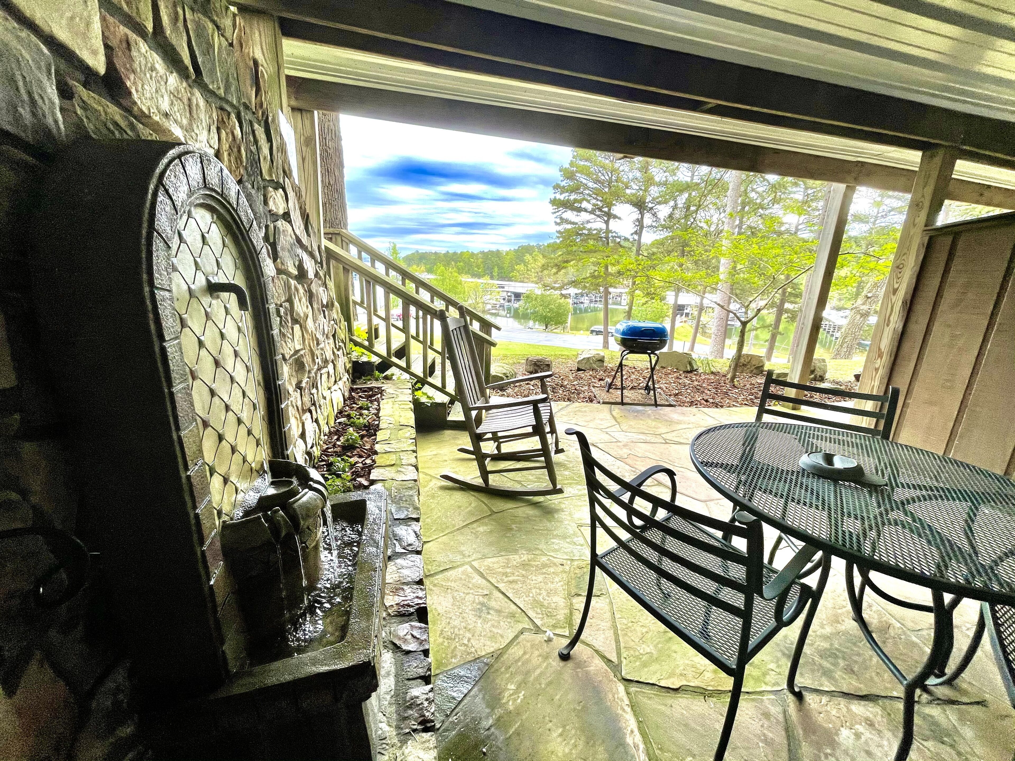 Un patio con chimenea de piedra, mobiliario exterior y vistas a un lago y árboles.