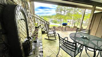 Un patio con chimenea de piedra, mobiliario exterior y vistas a un lago y árboles.