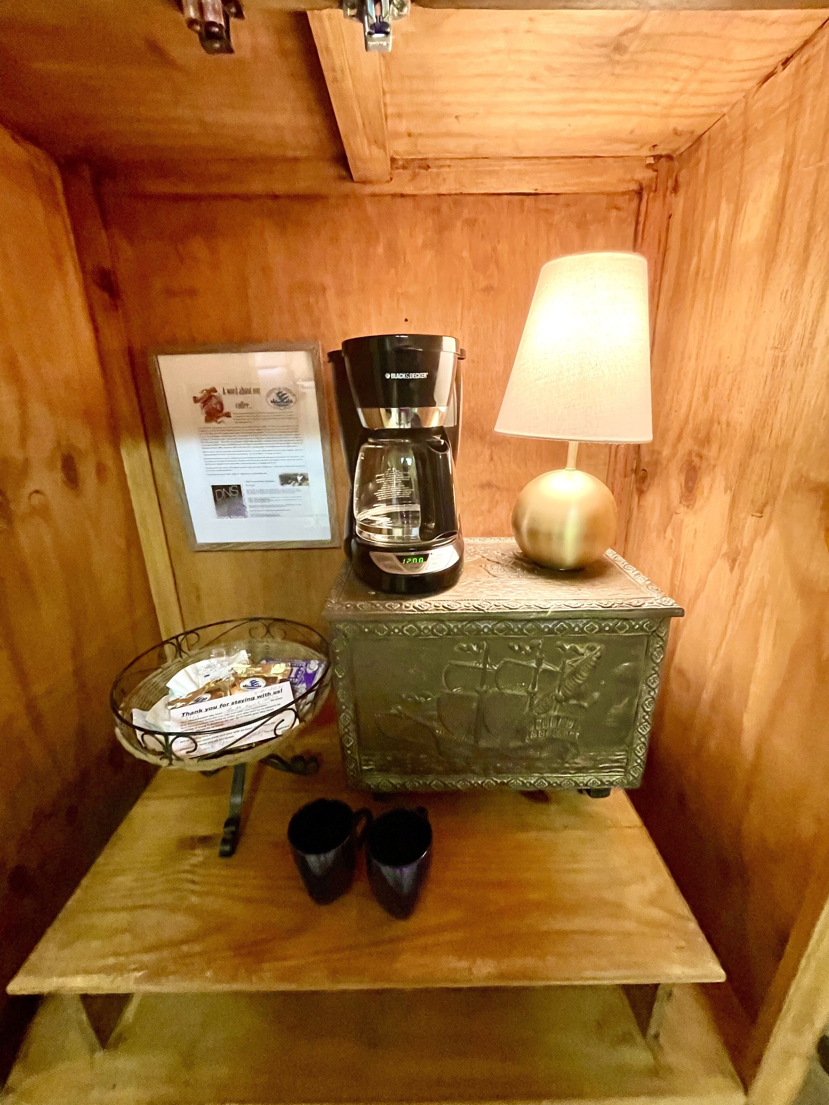 A small wooden room with a coffee maker, a lamp, a glass bowl, and a decorative box on a table.