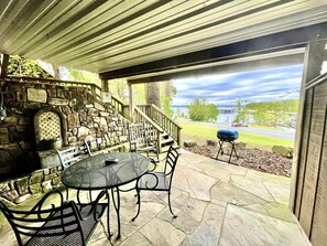 A covered patio with stone walls, a stone fireplace, outdoor seating, and a view of a lake and road. - SU15, Spacious studio with full kitchen, dining table and rock patio overlooking Lake Ouachita. by RedAwning (Mount Ida)