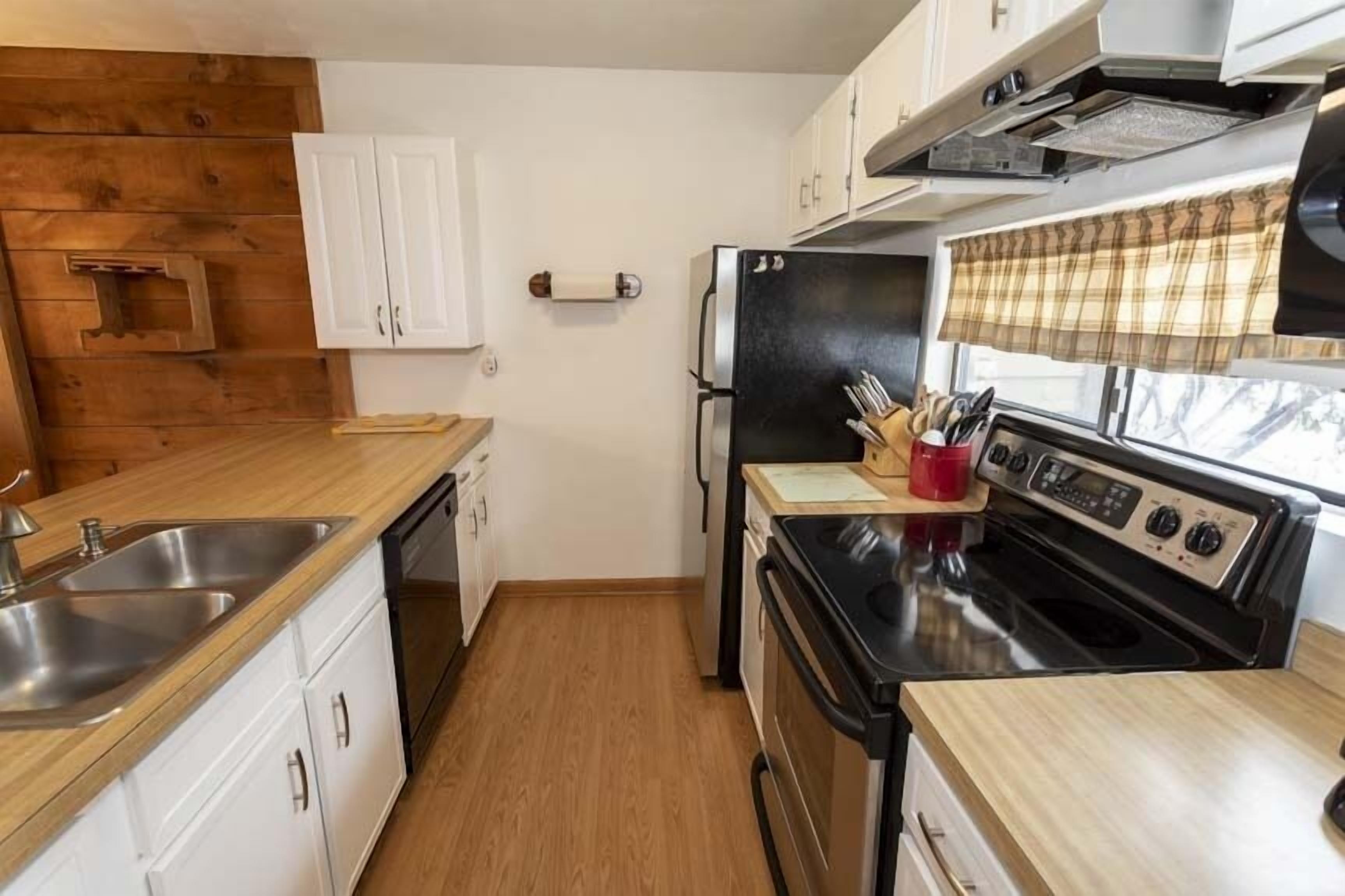 Une cuisine de taille réduite avec des meubles blancs, un réfrigérateur noir, une cuisinière et une fenêtre agrémentée d’un rideau à carreaux.