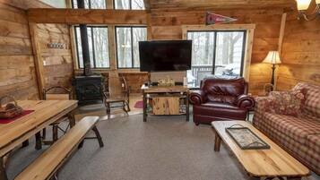 Een cabin interieur met houten wanden, een open haard, een televisie, een leren fauteuil, een bank en een eettafel met banken.