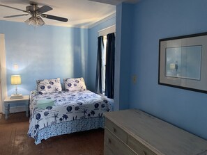 Un dormitorio con cama, mesita de noche, cómoda, ventilador de techo y ventana con cortinas.