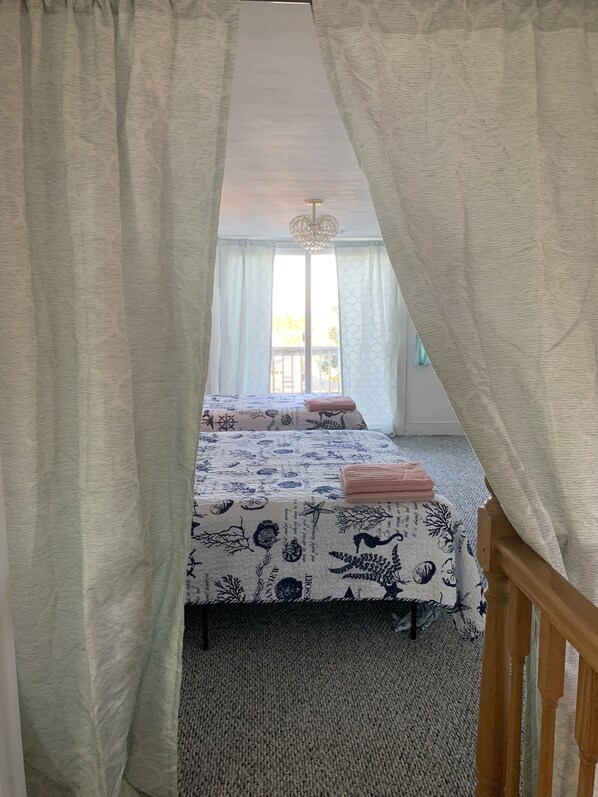 A bedroom with two beds, a wooden headboard, a large window with sheer curtains, and a chandelier. - Mermaids & Mutts 3 Bedroom Bungalow by RedAwning (Norfolk)