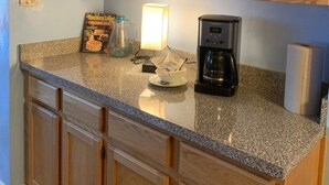 A kitchen with wooden cabinets, a granite countertop, a coffee maker, and a bowl with coffee pods. - Mermaids & Mutts 3 Bedroom Bungalow by RedAwning (Norfolk)