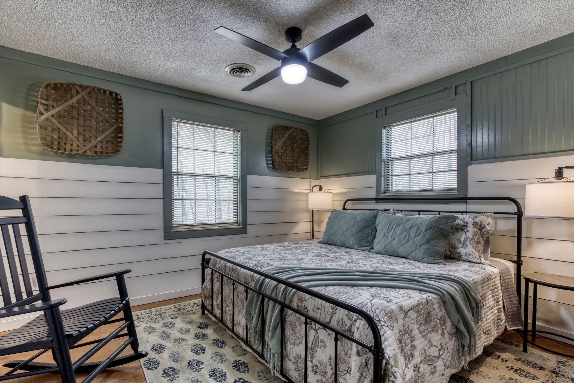 A bedroom with a bed, a chair, a ceiling fan, and two windows with blinds.