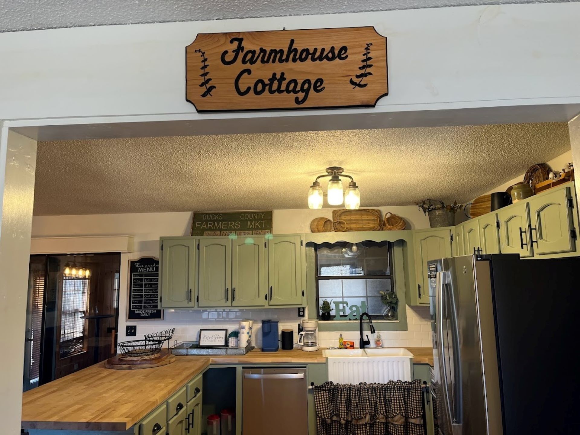 A kitchen with green cabinets, a farmhouse sign, a menu board, and a window with the word "Eat" on it.