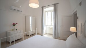 Quarto com cama, penteadeira, radiador, ar condicionado, janela com cortinas e porta.