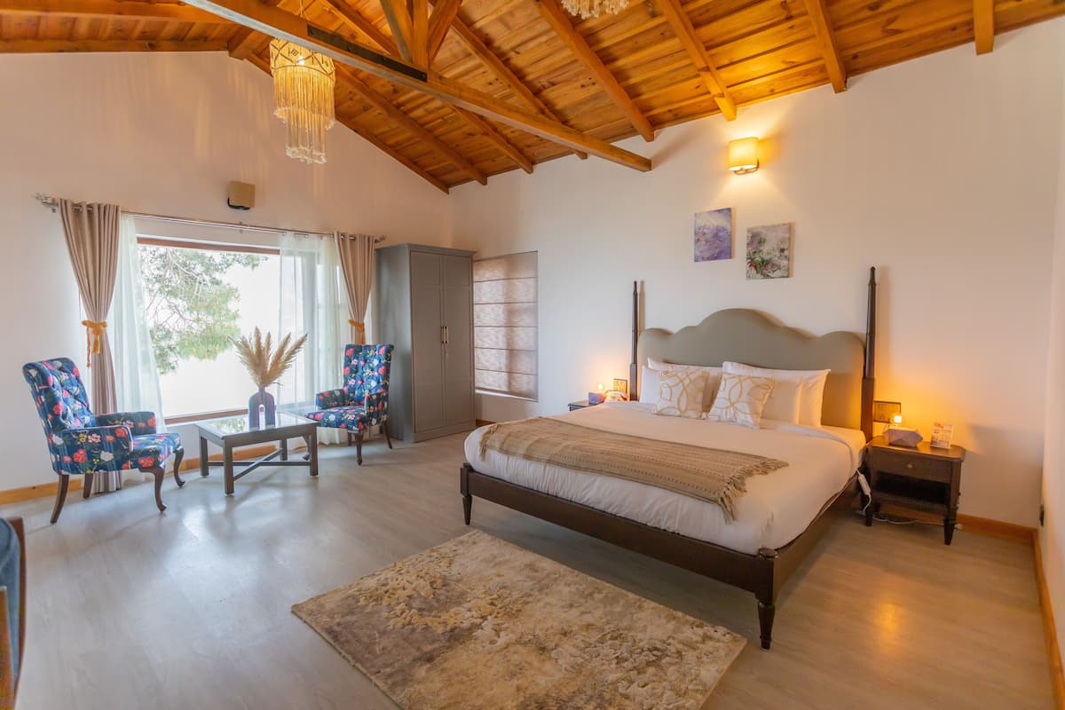 A bedroom with a large bed, a wooden ceiling, a nightstand, a chair, a table, and a window with curtains.