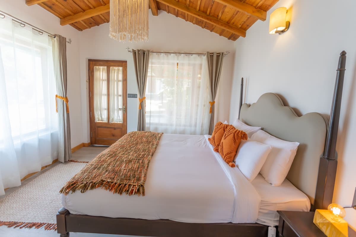A bedroom with a large bed, wooden ceiling beams, a chandelier, and a nightstand with a lamp.