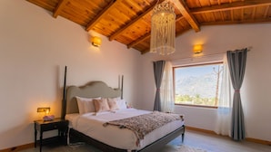 Een slaapkamer met een houten plafond, een groot bed met wit beddengoed, een nachtkastje met een lamp en uitzicht op bergen door een raam met gordijnen.
