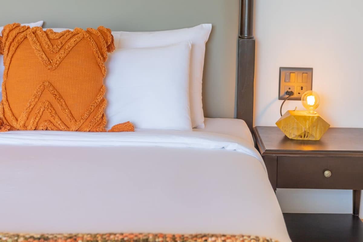 A neatly made bed with a white and orange pillow, a patterned throw blanket, and a bedside table with a lamp.