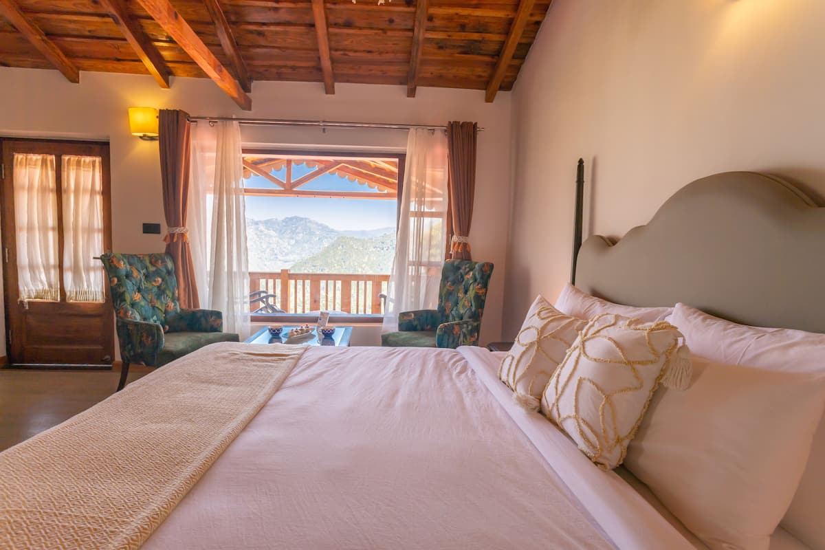 A bedroom with a large bed, two chairs, a wooden ceiling, a window with curtains, and a view of mountains.