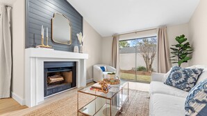 Un salon avec une cheminée, une table basse en verre, un canapé blanc et une grande fenêtre avec des rideaux.