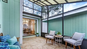 Un patio couvert aménagé avec des fauteuils en osier et un banc rembourré, une petite table ornée d’une plante, et une vue sur l’intérieur à travers une porte vitrée.