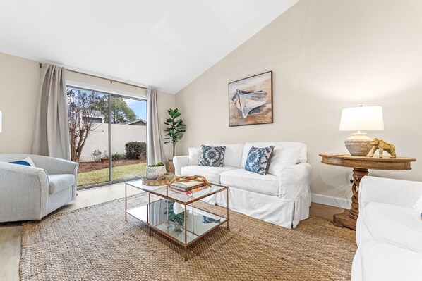 A modern living room with a glass coffee table, white sofas, a round wooden side table, and a painting on the wall. - Beach Villa w/ 4-Seat GOLF CART! Near Beach & Pool. Screened Porch, Yard (Miramar Beach)