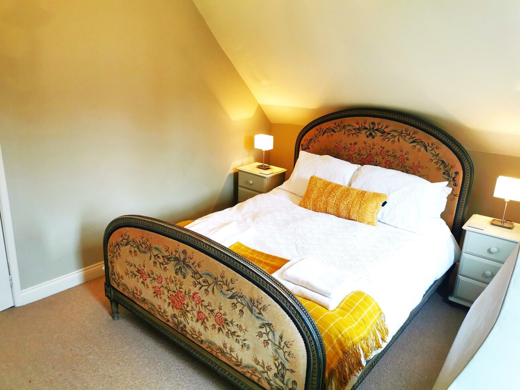 A neatly made bed with floral headboard and footboard, two bedside tables with lamps, and a carpeted floor.