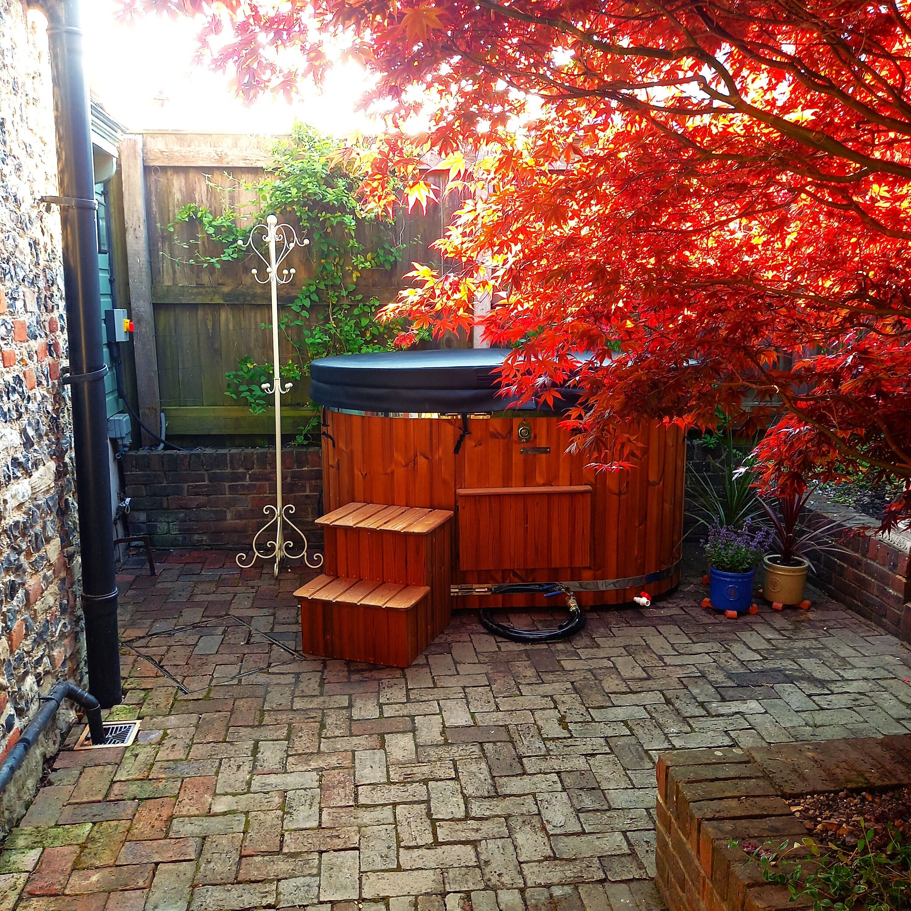 A wooden hot tub with steps, surrounded by a brick patio and red foliage.