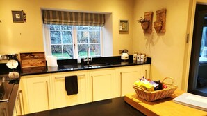A kitchen with a black countertop, a window with a striped valance, and a wooden cutting board with a basket of items. - Beautiful, traditional rural cottage (Shrewton)