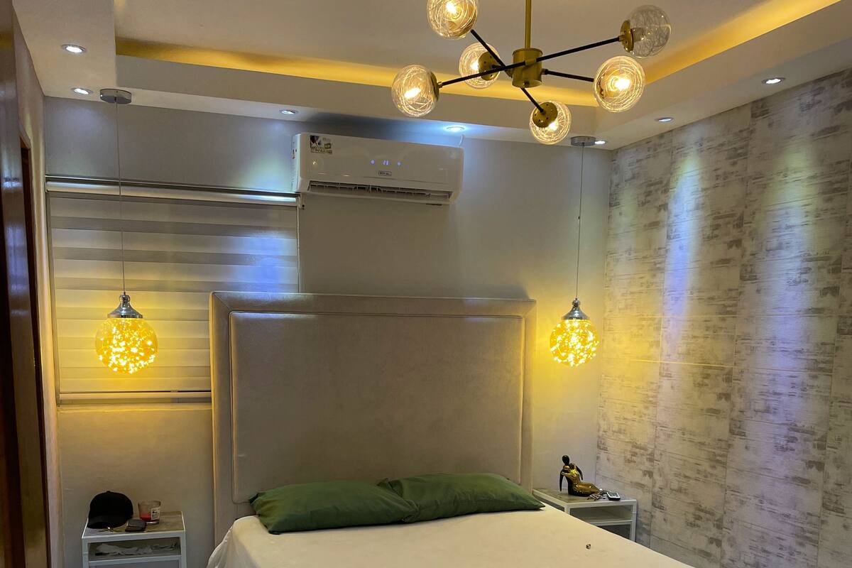 Une chambre moderne avec un grand lit, une table de chevet, un climatiseur fixé au mur et un luminaire au plafond au design original.