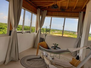 Une pièce avec un hamac, un tapis rond et une vue sur la verdure et les bâtiments.