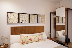 Camera d'albergo con una testiera in legno, un letto con lenzuola bianche, un cuscino floreale, un comodino con una lampada e una parete decorata con quattro quadri astratti.